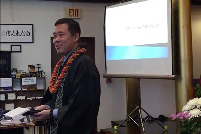 Blayne Higa, in robes and wearing lei, speaks at Moiliili Hongwanji